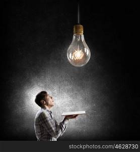 In search of bright inspiration. Young man holding opened book and glass glowing bulb hanging above