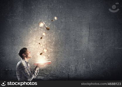 In search of bright inspiration. Young man holding opened book with glass glowing light bulbs flying out