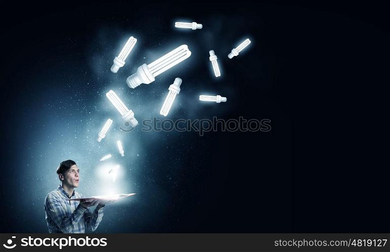 In search of bright inspiration. Young man holding opened book with glass glowing light bulbs flying out