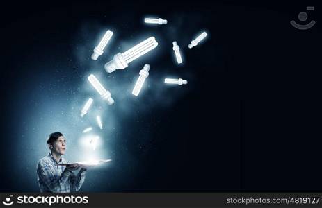 In search of bright inspiration. Young man holding opened book with glass glowing light bulbs flying out