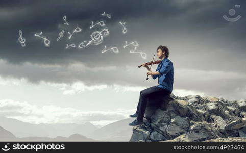 In search of inspiration. Young handsome man sitting on rock edge and playing violin