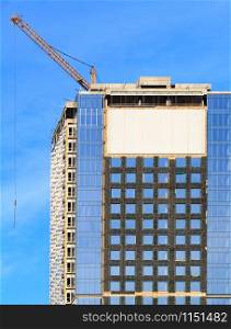 In the construction of modern residential concrete buildings with a glass facade, a tower crane is used, the blue sky is reflected in the windows of the building, vertical image with copy space.. Construction of a modern concrete building with a glass facade, a reflection of the blue sky and tower cranes.