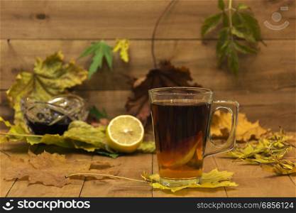 In the foreground is in focus glass cup of tea. In the background, the focus is visible without lemon, vase with jam and autumn leaves.
