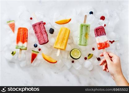 In the girl&rsquo;s hand, a healthy berry ice cream lolly . On the ice cubes, a different ice cream with pieces of fruit and fresh berries. Cold dessert. Flat lay. A woman&rsquo;s hand holds an ice cream popsicle against a background of ice cubes with ice cream and a piece of fruit. Flat lay