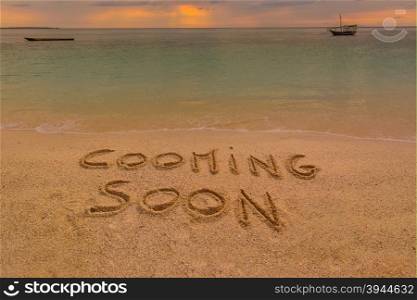 "In the picture a beach at sunset with the words on the sand "Cooming soon"."