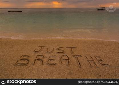 "In the picture a beach at sunset with the words on the sand "Just breathe"."