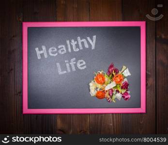 "In the picture a blackboard, on the left side with written "Healthy Life" and on the right side salad of vegetables on wooden background"