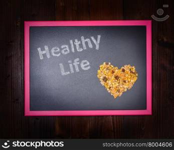 "In the picture a blackboard, on the left side with written "Healthy Life" and on the right side a mixture of cereals which draw a heart on wooden background"