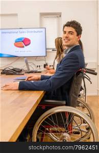 inclusion - portrait of a man in wheelchair. Inclusion - portrait of a man in a wheelchair participating in a meeting with colleagues in an office environment with a screen in the background showing international sales figures