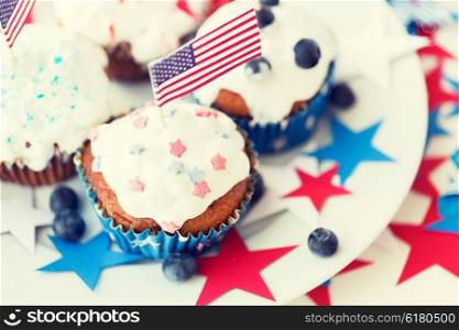 independence day, celebration, patriotism and holidays concept - close up of glazed cupcakes or muffins decorated with american flags and blueberries on plate at 4th july party