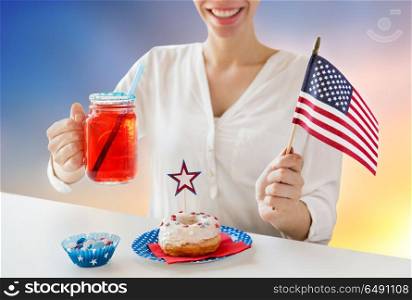 independence day, patriotism and holidays concept - close up of happy woman with donut celebrating 4th july, holding american flag and drinking juice from glass mason jar over evening sky background. happy woman celebrating american independence day. happy woman celebrating american independence day