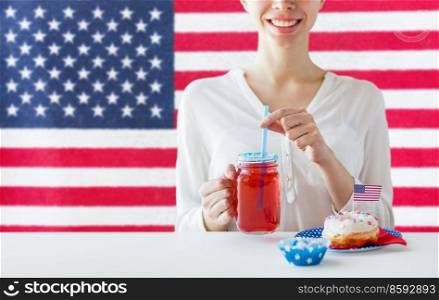 independence day, patriotism and holidays concept - close up of happy woman with iced donut drinking juice from mason jar glass at 4th july party over flag of united states of america on background. happy woman celebrating american independence day