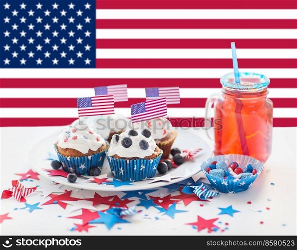 independence day, patriotism and holidays concept - close up of iced cupcakes, juice glass or mason jar and candies at 4th july party over flag of united states of america on background. cupcakes with american flags on independence day