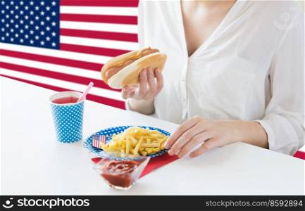 independence day, patriotism and holidays concept - close up of woman eating hot dog and french fries with drink in paper cup at 4th july at party over flag of united states of america on background. close up of woman eating hotdog and french fries