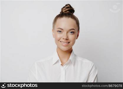 Independent business woman in white shirt with hair in bun feeling happy at work and being in good mood, female manager thinking about new progressive ideas, standing alone in front of light wall. Happy successful business woman in white shirt posing in studio
