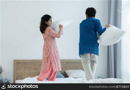 Indian family, brother and sister with traditional clothes having fun playing pillow fight, two children standing on bed together at bedroom at home. Sibling relationship concept