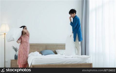Indian family, brother and sister with traditional clothes having fun playing pillow fight, two children standing on bed and floor at bedroom at home. Sibling relationship concept