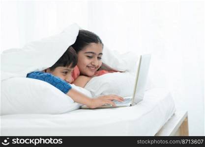 Indian family, brother and sister with traditional clothes smiling using laptop in bedroom at home, two children lying on bed and white pillow under blanket duvet. Sibling relationship concept