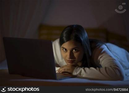 Indian girl looking at camera while lying on bed with laptop