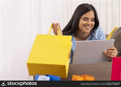 Indian girl watching tablet while lying on sofa with shopping bag in her hand