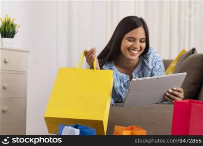 Indian girl watching tablet while lying on sofa with shopping bag in her hand