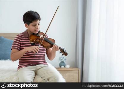 Indian handsome kid is actively practicing playing wooden violin at home. Happy Asian teenage boy learning violin instrument alone, sitting on bed by the window in bedroom. Education concept