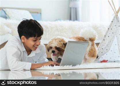 Indian handsome teenage boy lying on floor using laptop computer, relaxing playing with shih tzu dog puppy in bedroom at home. Pet friendly residence, lifestyle, pet and human bonding concept