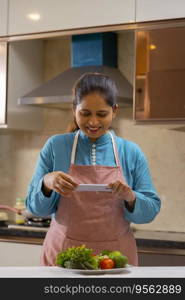 Indian woman taking photograph of vegetables with her mobile in the kitchen