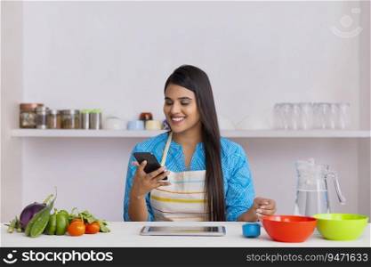 Indian woman watching smartphone while sitting in kitchen