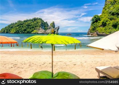 Indonesia. A small sandy beach on a tropical island. Umbrellas and sunbeds in the foreground. Ocean and rocky islet in the background. Nobody. Umbrellas and Sunbeds on a Tropical Beach