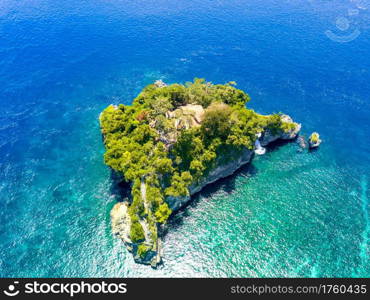 Indonesia. Emerald ocean water. A small rocky island, overgrown with jungles. Stairs from the water and a few huts between the trees. Aerial view. Small Rocky Island in the Ocean and Huts. Aerial View