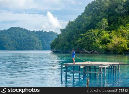 Indonesia. Rustic pier on one of the thousands of tropical islands. The boy puts the bait on the hook of his fishing rod. Boy Fisherman on the Rustic Pier of a Tropical Island