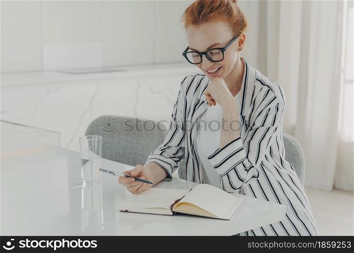 Indoor image of happy ginger business woman dressed casually sitting at table in kitchen, making notes and drinking pure water from glass in morning while planning her day, writing down thoughts ideas. Happy ginger woman taking notes while planning her day in morning at home, drinking pure water