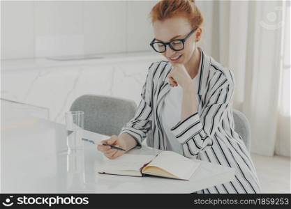 Indoor image of happy ginger business woman dressed casually sitting at table in kitchen, making notes and drinking pure water from glass in morning while planning her day, writing down thoughts ideas. Happy ginger woman taking notes while planning her day in morning at home, drinking pure water