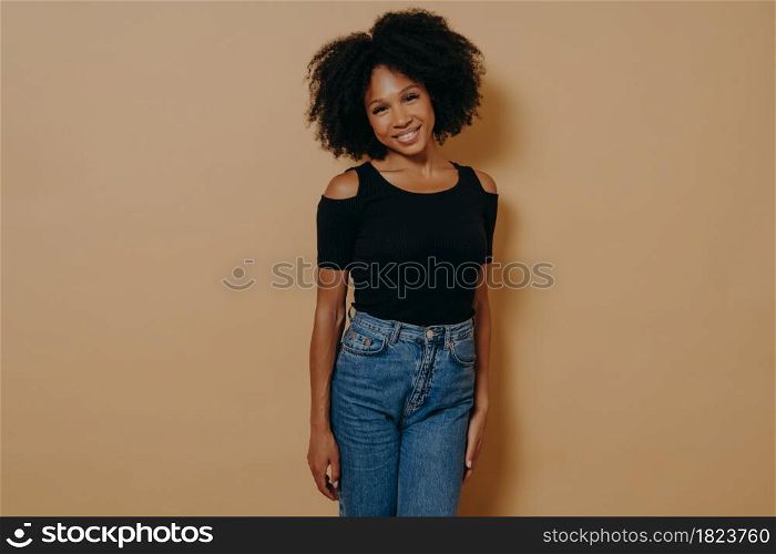 Indoor photo of pretty afro american smiling woman in casual outfit looking at camera with shy charming smile, posing isolated over dark beige background. Beautiful dark skinned women. Pretty afro american woman looking at camera with shy charming smile againt dark beige wall