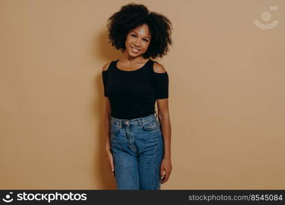 Indoor photo of pretty afro american smiling woman in casual outfit looking at camera with shy charming smile, posing isolated over dark beige background. Beautiful dark skinned women. Pretty afro american woman looking at camera with shy charming smile againt dark beige wall