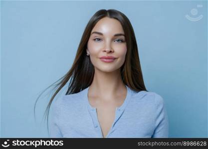 Indoor portrait of pretty woman feels happy and carefree during day off, has long straight hair, wears blue jumper, enjoys positive conversation, stands indoor. Women, lifestyle and well being concept