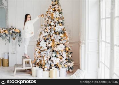 Indoor shot of adorable little kid stands on chair on tiptoes, decorates big New Year tree, has good mood, being in anticipation of holidays and wants to recieve presents. Celebration and decoration