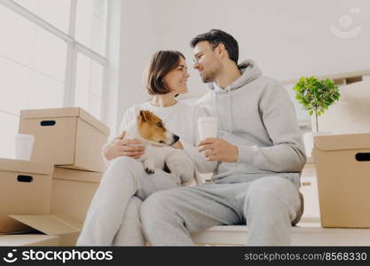 Indoor shot of affectionate woman and man express love to each other, have good relationship, drink coffee, pose with favourite pet, have to unpack many cardboard boxes, bought new apartment