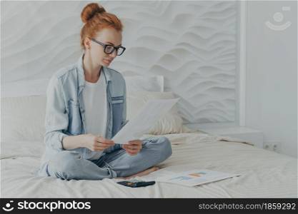 Indoor shot of attractive redhead young woman sits crossed legs on comfortable bed checks financial documents calculates family budget or household expenditures wears denim clothes being at home. Young woman sits crossed legs on comfortable bed checks financial documents calculates family budget