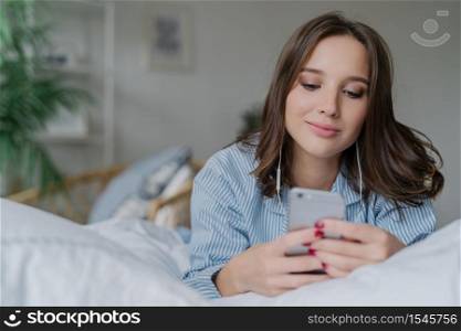 Indoor shot of attractive thoughtful young woman lies in bed, uses modern cell phone, watches video or film online, enjoys good sound, dressed in pyjamas. People, bed time and relaxation concept