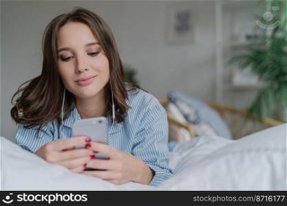 Indoor shot of attractive thoughtful young woman lies in bed, uses modern cell phone, watches video or film online, enjoys good sound, dressed in pyjamas. People, bed time and relaxation concept