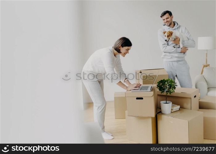 Indoor shot of attractive woman leans to laptop computer, focused into screen, surfes internet, searches ideas for decoration, man stands near, plays with favourite pet, move into new house.