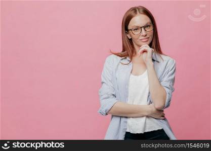 Indoor shot of attractive young lady keeps hands partly crossed, looks seriously at camera, wears elegant shirt and trousers, models over pink studio wall with copy space for your advertisement