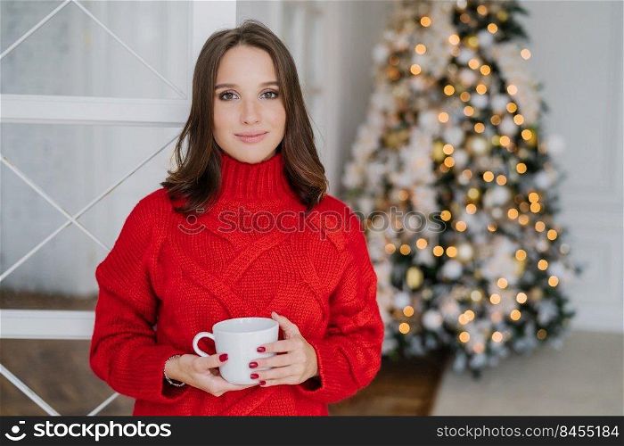 Indoor shot of beautiful Caucasian woman dressed in war  pullover, drinks hot beverage, enjoys domestic atmopshere, prepares for celebrating Christmas or New Year. People and lifestyle concept