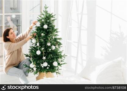 Indoor shot of beautiful happy woman in winter sweater and jeans decorates Christmas tree, celebrates Ney Year eve, spends time at home, poses in bedroom with white walls. Holiday celebration