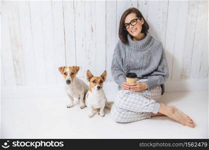 Indoor shot of beautiful woman wears spectacles and casual wear, drinks hot beverage, enjoys good morning, sits near her favourite pets against white fence background. Happiness and leisure concept