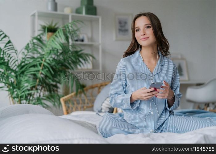 Indoor shot of beautiful young woman watches favourite serial with earphones and smart phone, downloads new songs, looks pensively aside, sits on bed in cozy room. Bed time and leisure concept