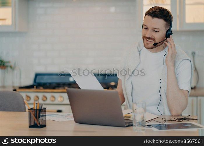 Indoor shot of busy European man has video courses watches webinar listens lecture studies online wears headphones focused at laptop screen dressed casually poses against cozy home interior.