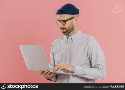 Indoor shot of busy male freelancer wears optical glasses for good vision, holds modern electronic gadget, surfes internet, develops new website, isolated over pink background. Technology concept